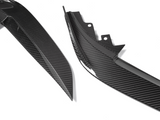 Two carbon fiber car body parts lie on a white surface, showcasing sleek, angular designs and the distinctive checkered pattern typical of carbon fiber material.