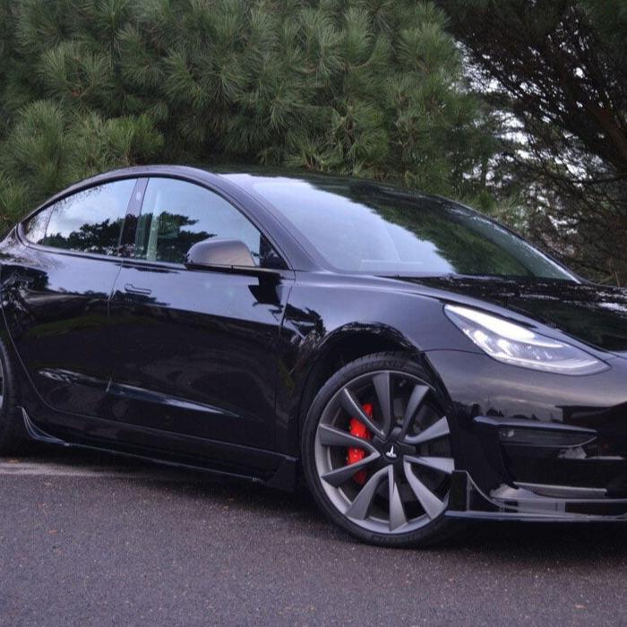 A black car is parked on asphalt, featuring red brake calipers and dark alloy wheels, with pine trees in the background.