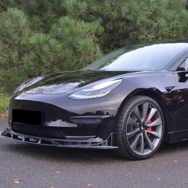 A sleek black car with silver alloy wheels is parked on a road, surrounded by dense green foliage in the background.