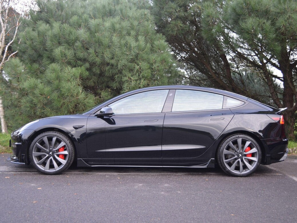 A black sedan is parked on a paved road, featuring red brake calipers and large wheels. It is positioned in front of dense green trees.