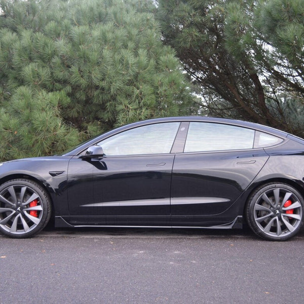 A black sedan is parked on a paved road, featuring red brake calipers and large wheels. It is positioned in front of dense green trees.