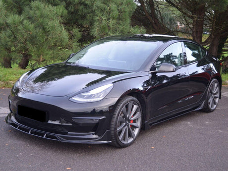 A sleek black car is parked on a paved area, surrounded by lush green trees, showcasing its streamlined, modern design and alloy wheels.