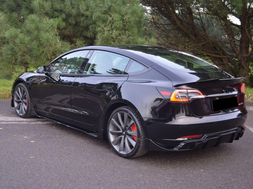 A sleek black car is parked on a paved road, surrounded by lush green trees, with sunlight reflecting off its polished surface.