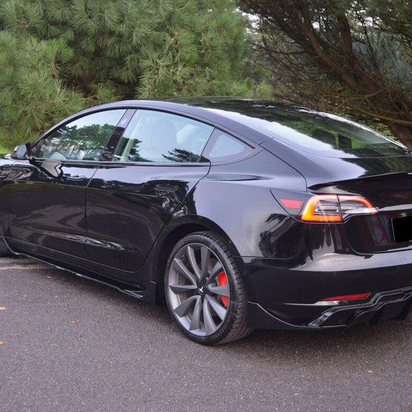 A sleek black car is parked on a paved road, surrounded by lush green trees, with sunlight reflecting off its polished surface.