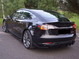 A sleek black electric car is parked on an asphalt surface, surrounded by lush green trees, with reflections visible on its glossy finish.