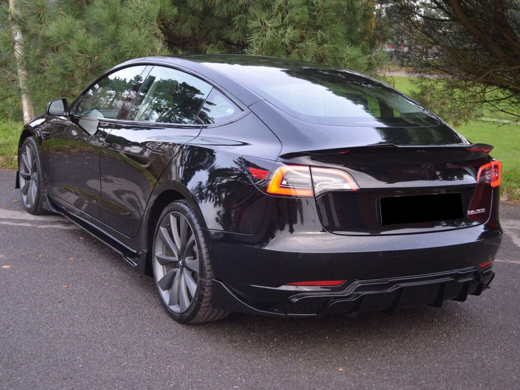 A sleek black electric car is parked on an asphalt surface, surrounded by lush green trees, with reflections visible on its glossy finish.