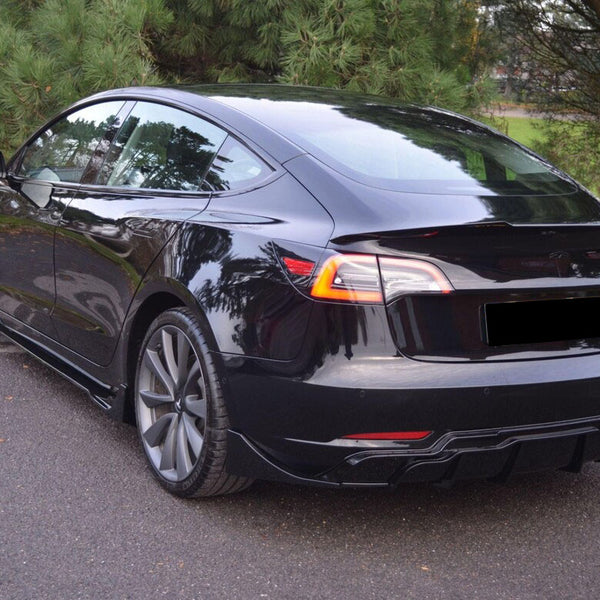 A sleek black electric car is parked on an asphalt surface, surrounded by lush green trees, with reflections visible on its glossy finish.