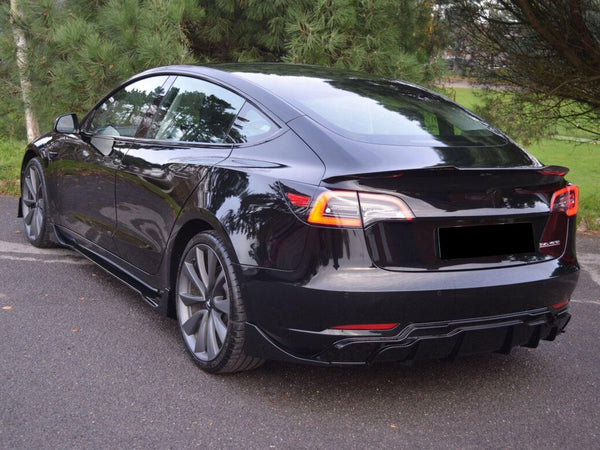 A sleek black electric car is parked on an asphalt surface, surrounded by lush green trees, with reflections visible on its glossy finish.