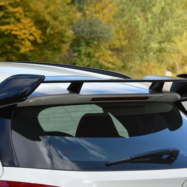 A sleek car spoiler perched on the rear of a white vehicle, situated against a backdrop of green and yellow foliage on a clear day.