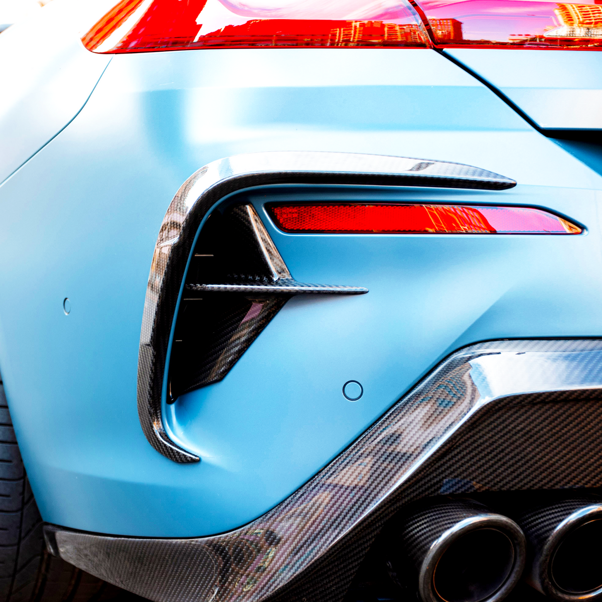 A blue sports car with a carbon fiber rear bumper features sleek, dual exhaust pipes and vibrant red taillights, parked outdoors.