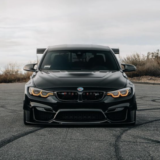 A sleek black sports car with glowing headlights is parked on an open asphalt road, surrounded by dry brush under a cloudy sky.