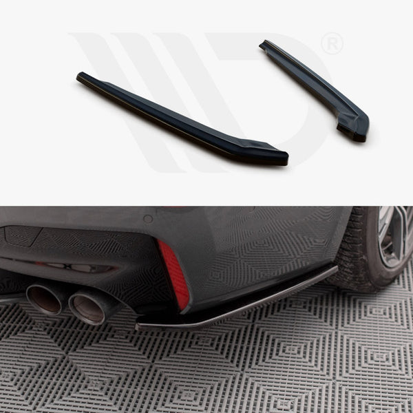 Two black car parts displayed above an installed piece on a car's rear bumper near dual exhaust pipes. The car is parked on a patterned surface.