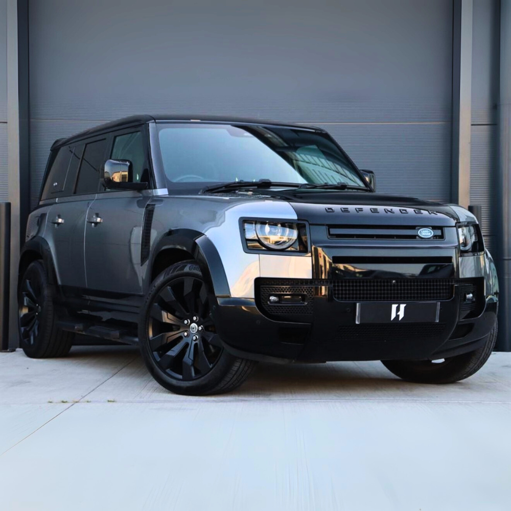 A grey Land Rover Defender with large black wheels is parked on a concrete surface in front of a closed grey garage door. "DEFENDER" is visible on the vehicle‚Äôs front.