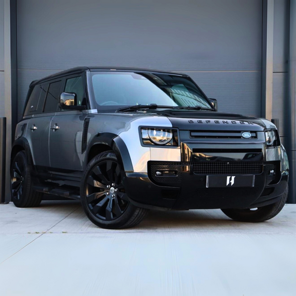 A grey Land Rover Defender with large black wheels is parked on a concrete surface in front of a closed grey garage door. "DEFENDER" is visible on the vehicle’s front.