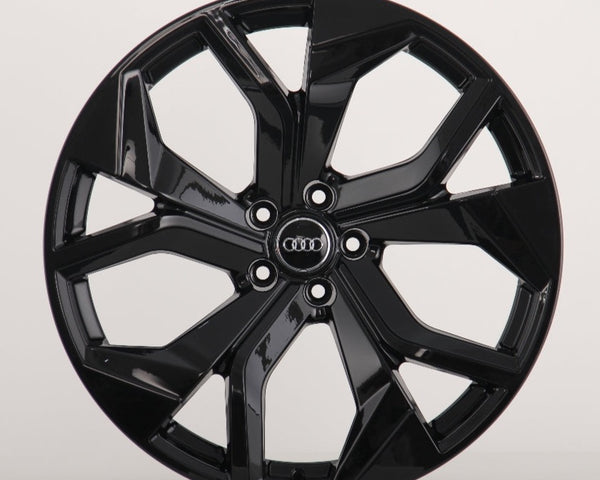 A glossy black car wheel rim with a modern design features five split spokes. In the center, four interlocking rings in silver logo represent Audi, against a plain white background.