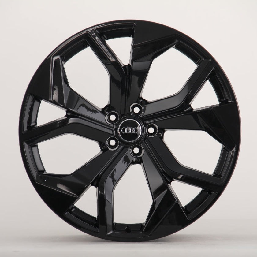 A glossy black car wheel rim with a modern design features five split spokes. In the center, four interlocking rings in silver logo represent Audi, against a plain white background.