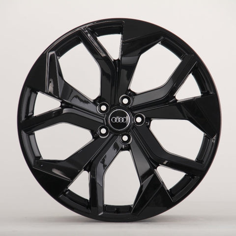A glossy black car wheel rim with a modern design features five split spokes. In the center, four interlocking rings in silver logo represent Audi, against a plain white background.