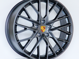 A dark alloy wheel with a multi-spoke design and a central Porsche logo is displayed upright against a plain white background.