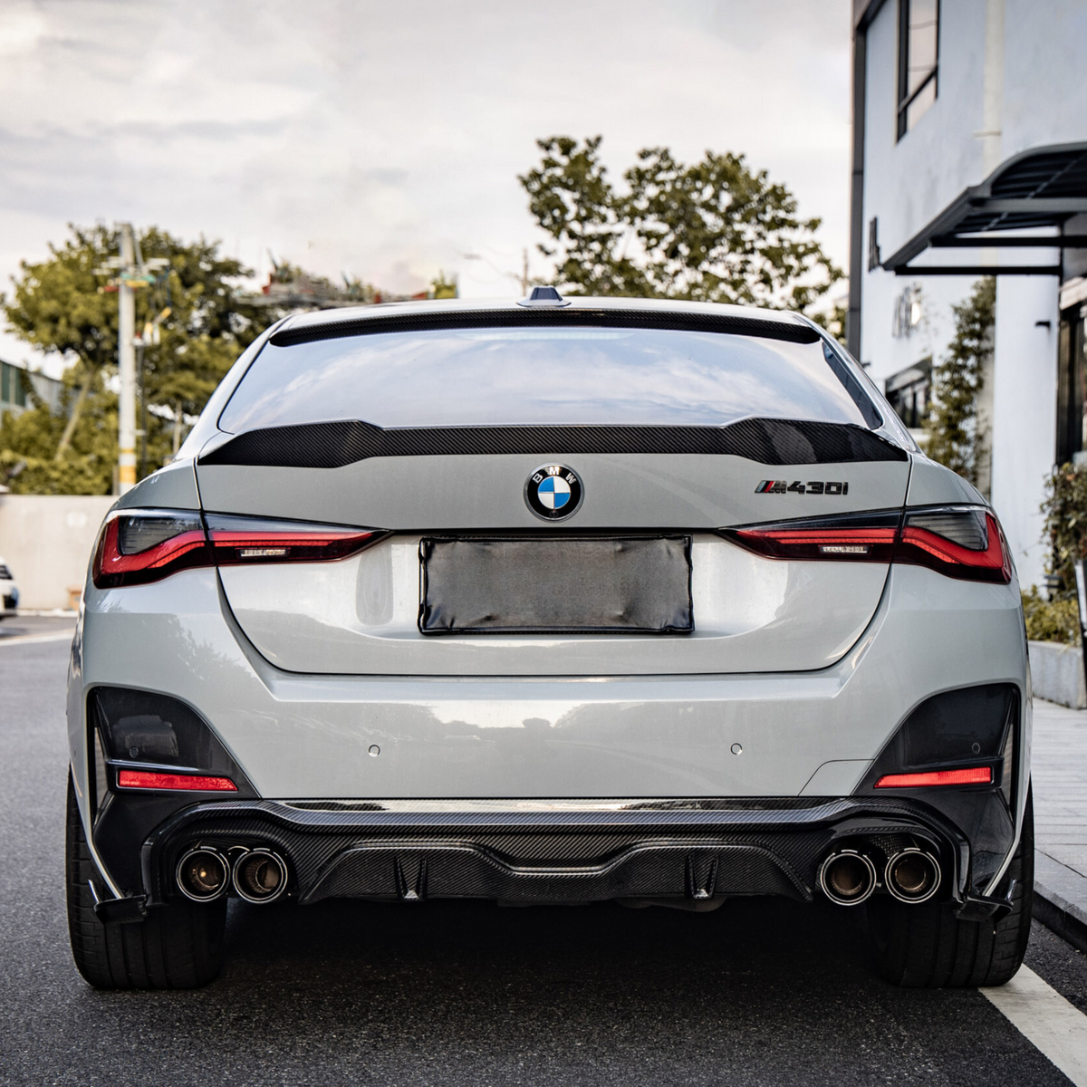 A silver BMW 4 Series (M440i) car is parked on a street, featuring dual exhaust pipes and a blurred license plate. Surrounded by trees and buildings under a cloudy sky.