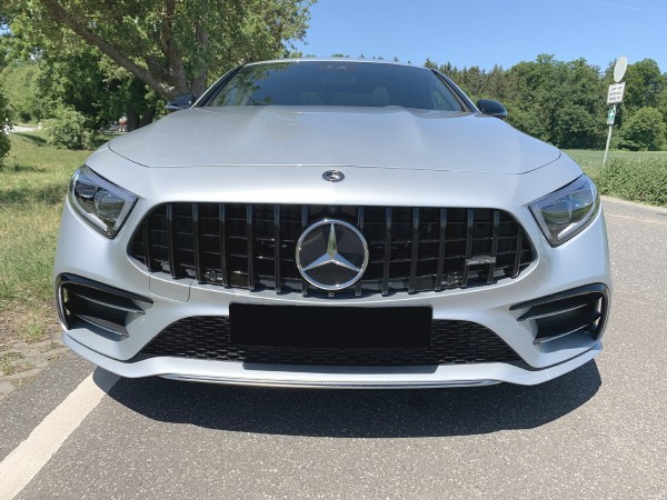 A silver Mercedes-Benz car is parked on a roadside, facing forward. It is under a clear blue sky, with green trees and grass in the background.
