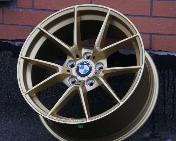 A gold BMW car wheel rim rests upright on a dark surface against a brick wall, displaying the BMW logo at its center.