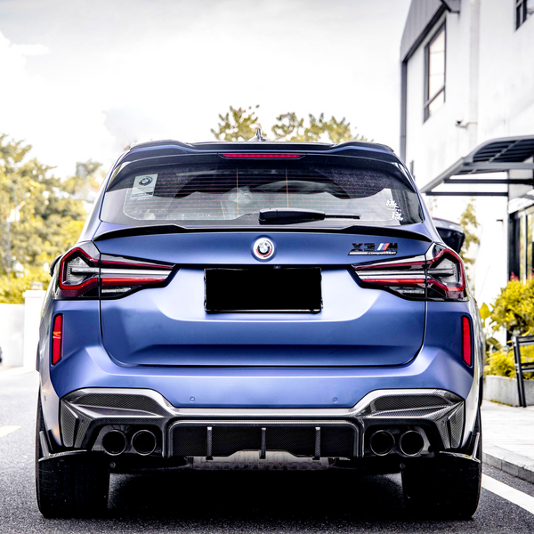 A blue BMW X3 M with a license plate concealed, parked outdoors. The badges on the back indicate “X3 M Competition.” Nearby are buildings and greenery.