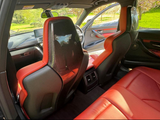 Rear view of a car interior features red leather seats with carbon fiber panels. The car door is open, revealing a grassy, sunlit park outside through the windows.