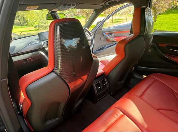 Rear view of a car interior features red leather seats with carbon fiber panels. The car door is open, revealing a grassy, sunlit park outside through the windows.