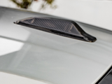 A carbon fiber vent is mounted on a smooth, light-colored car hood, situated in an outdoor setting with blurred foliage visible in the background.