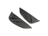 Two black carbon-fiber automotive splitters, each with angular shapes and grooves, are placed on a plain white surface, suggesting high-performance vehicle components.