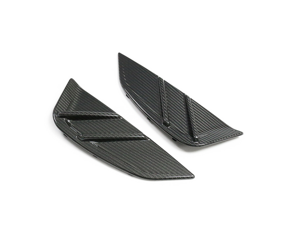 Two black carbon-fiber automotive splitters, each with angular shapes and grooves, are placed on a plain white surface, suggesting high-performance vehicle components.