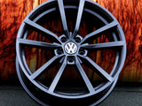 A black alloy wheel with the Volkswagen logo is propped against a textured, rust-colored background, resting on a dark asphalt surface.