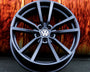 A black alloy wheel with the Volkswagen logo is propped against a textured, rust-colored background, resting on a dark asphalt surface.
