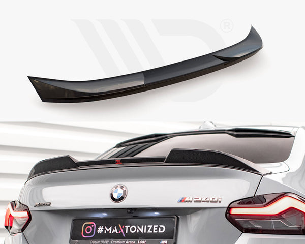 A glossy black rear spoiler sits unattached above a grey BMW M240i in a showroom. Text on the license plate reads "#MAXTONIZED" with an Instagram logo.