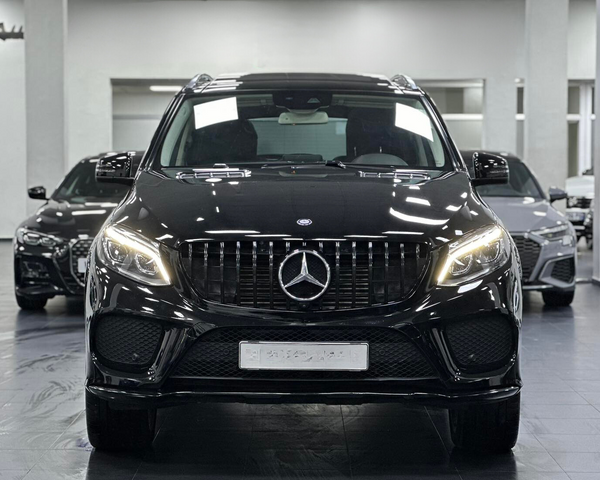A black Mercedes-Benz SUV stands prominently with headlights on, inside a showroom, flanked by two other black cars in the background.