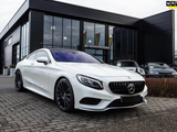 A white luxury car is parked outside a modern glass-walled showroom, positioned on a cobblestone driveway. The word 