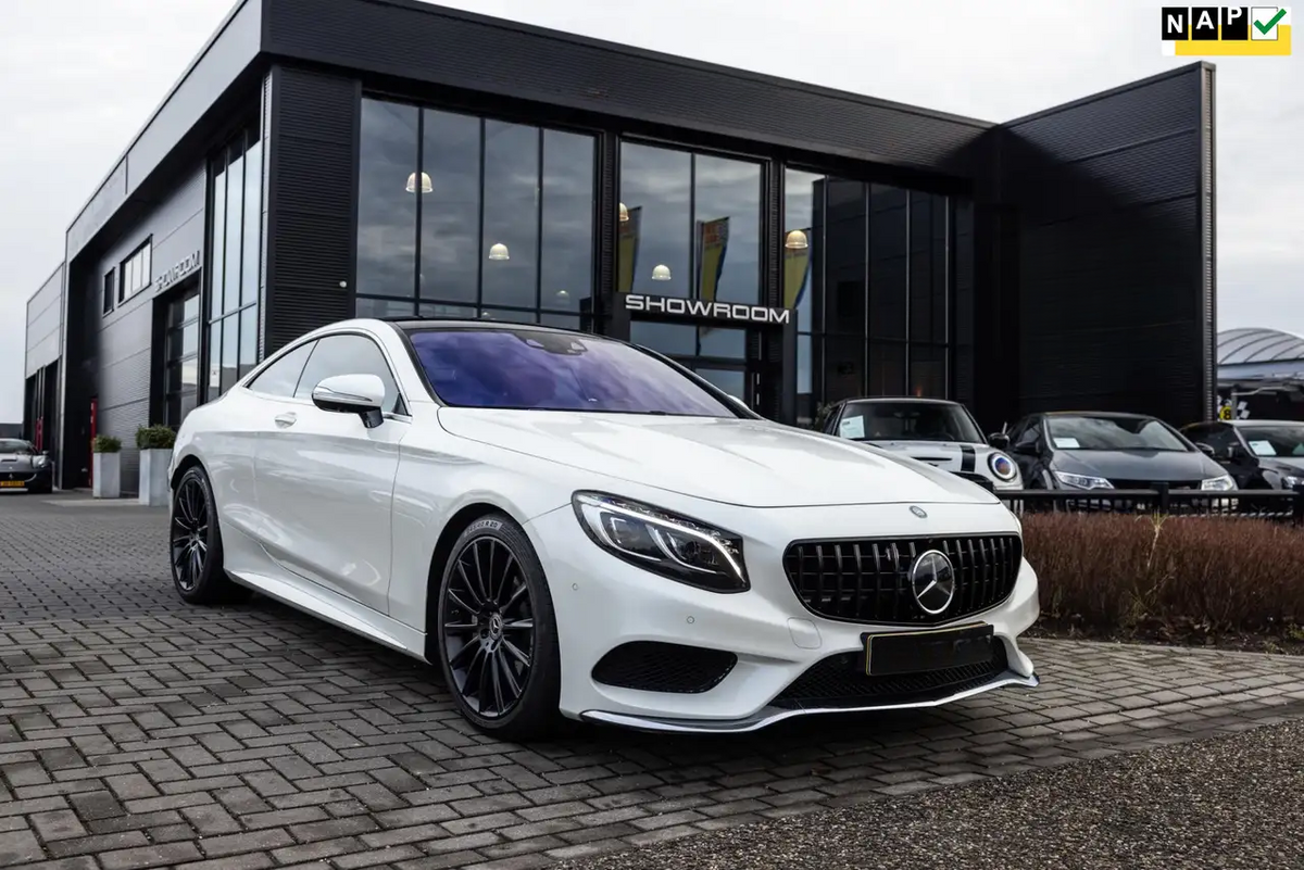 A white luxury car is parked outside a modern glass-walled showroom, positioned on a cobblestone driveway. The word "SHOWROOM" is visible on the building behind.