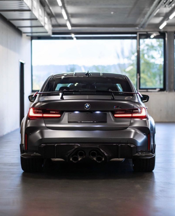 A sleek, dark-colored BMW car parked indoors with quad exhaust pipes, illuminated taillights, and a rear spoiler. Large windows reveal a blurred view outside.