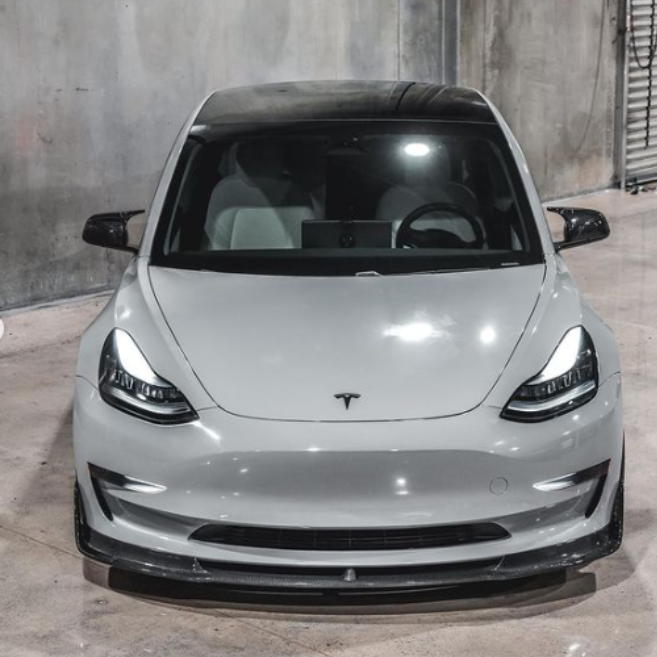 A sleek white car with a glossy finish is parked in an industrial garage with concrete walls. The front features a subtle emblem on the hood.