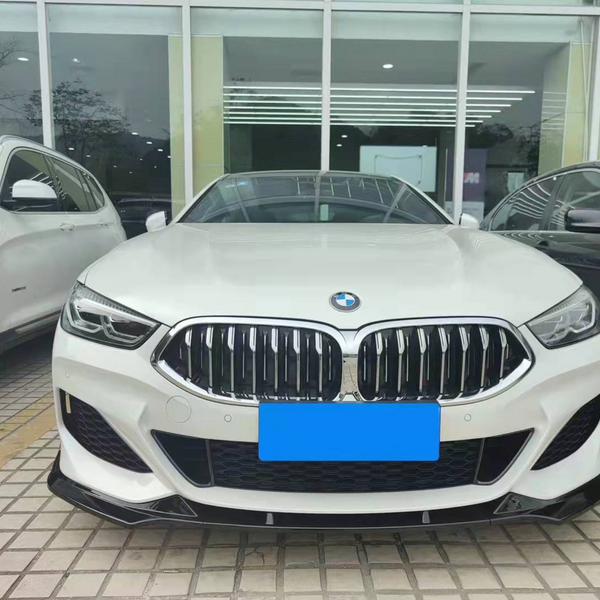 White BMW parked between two vehicles in front of a glass-fronted building. The car's front grille and headlights are prominently displayed on a tiled surface.