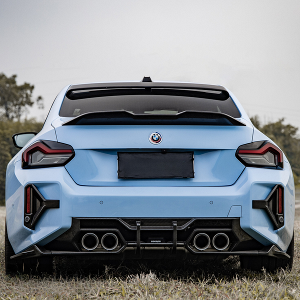 A light blue sports car is parked in a grassy area, displaying a sleek rear design with dual exhaust pipes and a visible logo above the license plate. Trees are in the background.