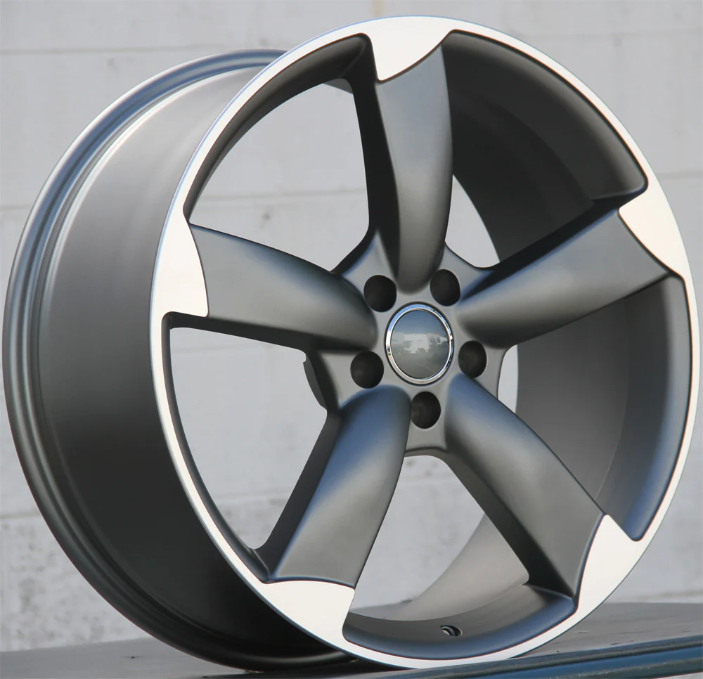 A metallic car wheel rim with five spokes, partially reflective, is propped upright against a plain, light-colored wall. The design features a combination of matte and glossy finishes.
