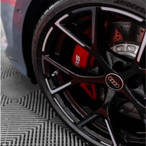 A black car wheel with an Audi logo is stationary, revealing a red brake caliper labeled "RS" in a garage setting with a checkered floor pattern.