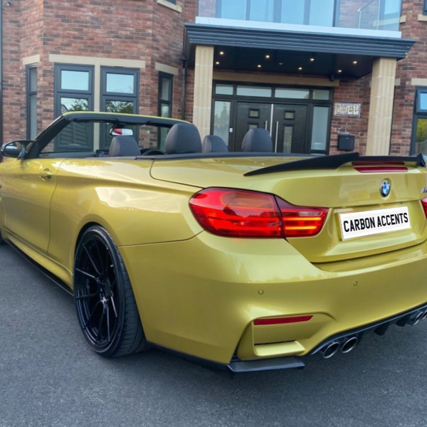 Convertible gold car with black wheels parked in front of a brick house with large windows and a double-door entrance. License plate reads "CARBON ACCENTS."