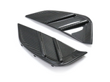 Two black carbon fiber car vents, angled and textured, with horizontal ridges. They rest on a plain white background, emphasizing their sleek, modern design.