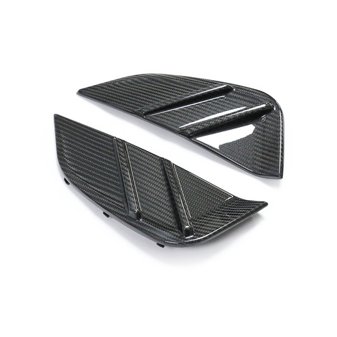 Two black carbon fiber car vents, angled and textured, with horizontal ridges. They rest on a plain white background, emphasizing their sleek, modern design.