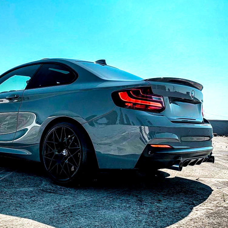 A gray sports car is parked on a sunlit concrete surface, with a clear blue sky overhead. The car's taillights are illuminated, emphasizing its sleek, aerodynamic design.