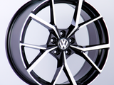 A black and silver alloy wheel with a Volkswagen logo in the center is displayed against a plain white background, highlighting the wheel's modern, geometric spoke design.