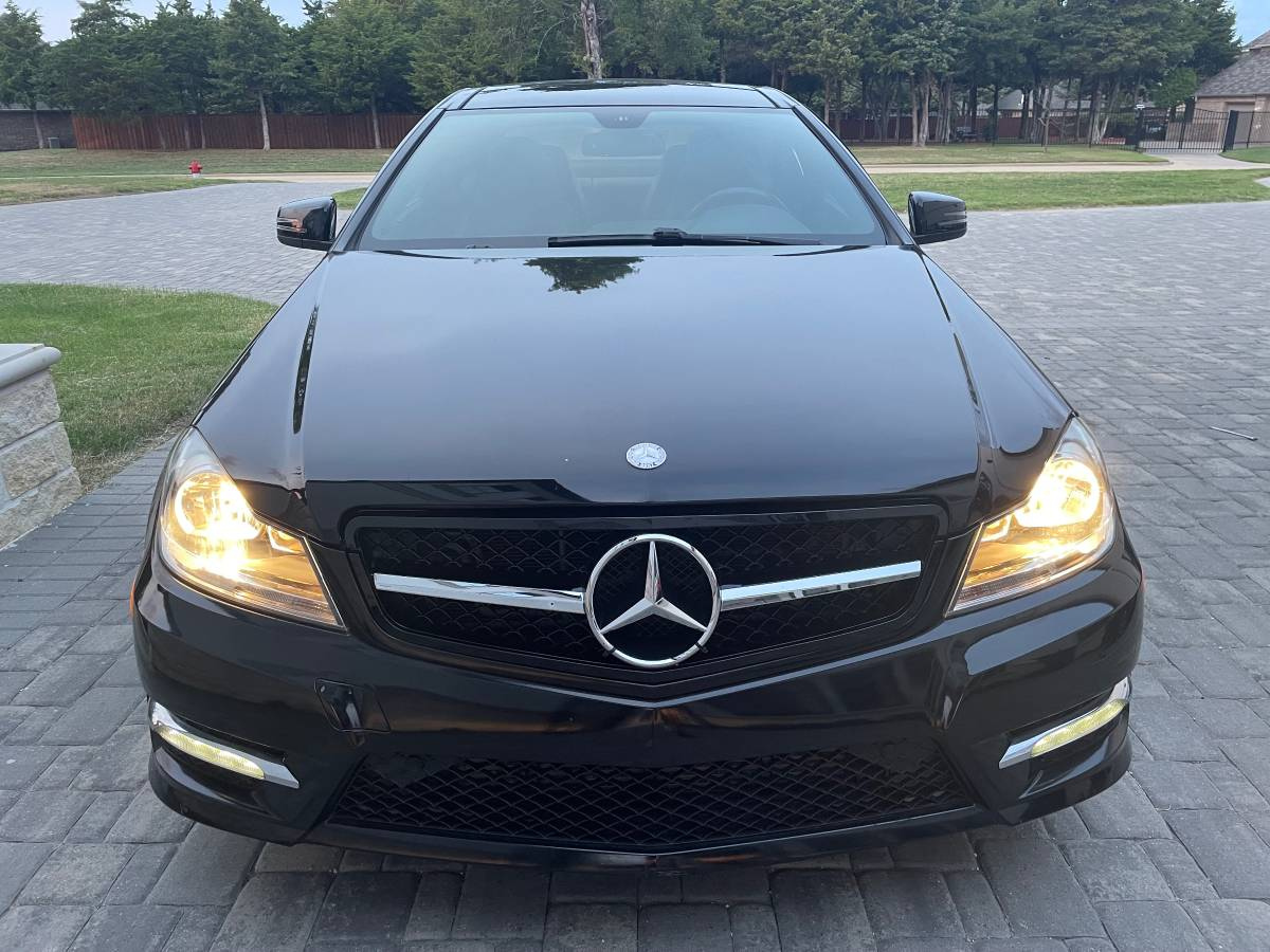 A black sedan with headlights on, parked on a paved driveway. It features a prominent emblem on the grille, surrounded by a residential area with trees and grass.