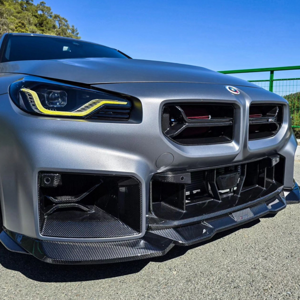 A sleek car with aggressive headlights and a carbon fiber front bumper is parked on asphalt, under a clear blue sky, with green railing and trees in the background.